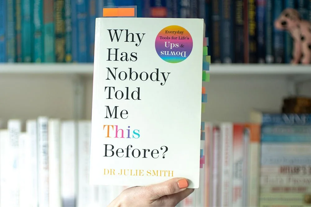 A copy of Why Has Nobody Told Me This Before? held up in front of a bookshelf